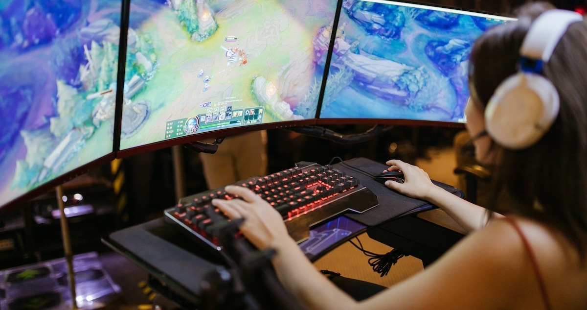 A female gamer with a headset seated at a desk in front of three gaming monitors. A visual representation of how to make a video game.